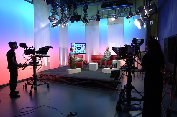 Blick in ein Fernsehstudio mit 2 Kameras, einer roten Couchgarnitur und zwei Talkgäste