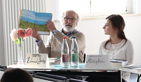 Ein Mann und eine Frau sitzen am Tisch, der Mann hält ein Buch hoch