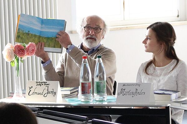 Ein Mann und eine Frau sitzen am Tisch, der Mann hält ein Buch hoch