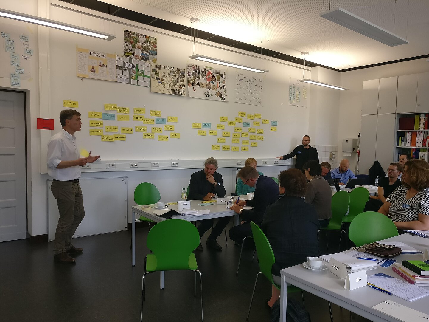 An einer Wand sind kleine gelbe und blaue Zettel geklebt. Ein Dozent spricht vor etwa einem Dutzend im Seminarraum befindlicher Teilnehmer.