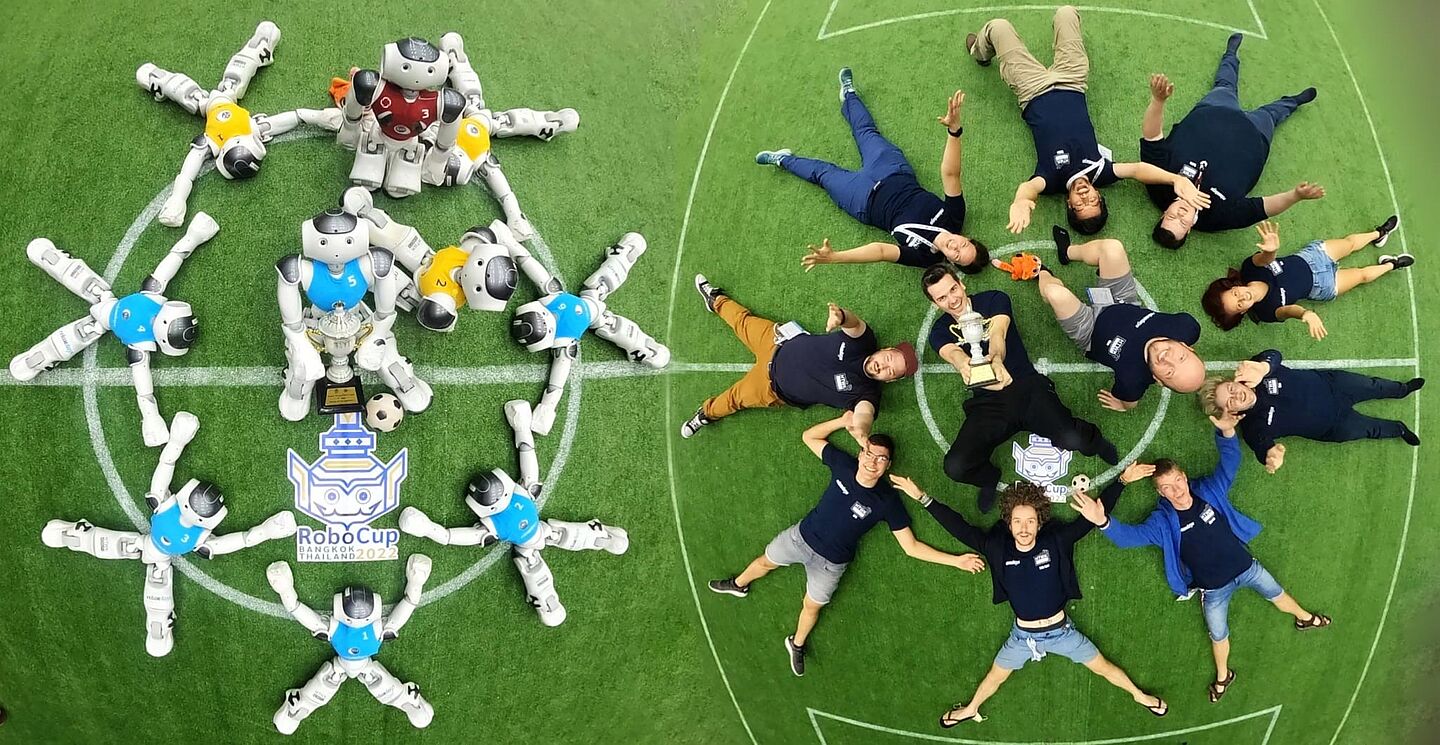 Die HTWK-Athletinnen und -Athleten – Vizeweltmeister der RoboCup-WM 2022 in Bangkok. Menschen und Roboter liegen auf grünem Grund und halten Pokal hoch