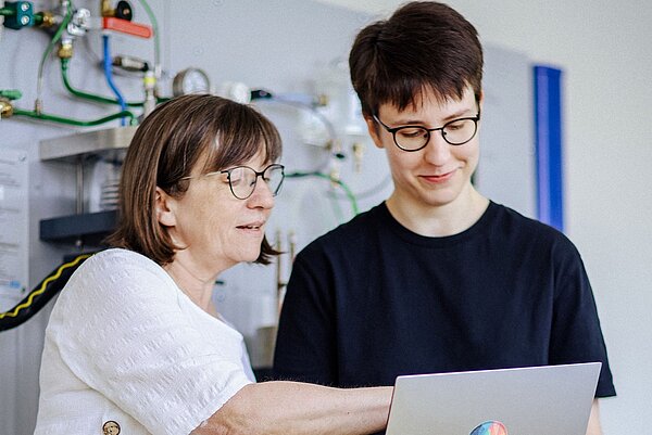 Zwei Personen stehen vor einer technischen Wandinstallation. Eine zeigt auf den Laptop der anderen, beide tragen Brillen und wirken konzentriert.