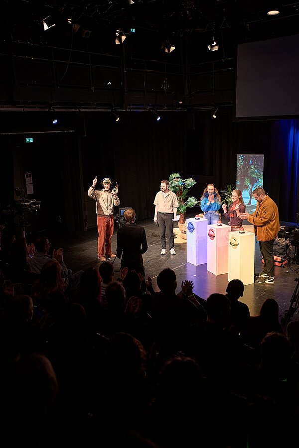 Es ist eine freudige Momentaufnahme des Spiels "Hornduras" zu sehen. Teilnehmende und der Spezialgast "Posthorn Paul" sind auf der Bühne zu sehen.