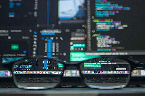 Zentrum digitale Arbeit (ZdA) Brille liegt auf einem Tisch vor zwei Bildschirmen, die verschiedene Programme zeigen