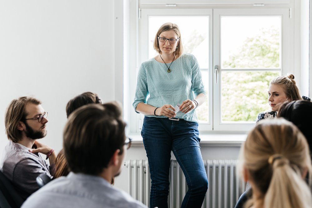 Mehrere Studierende sitzen vor einer lehrenden Frau, die entspannt vor ihnen steht und spricht.