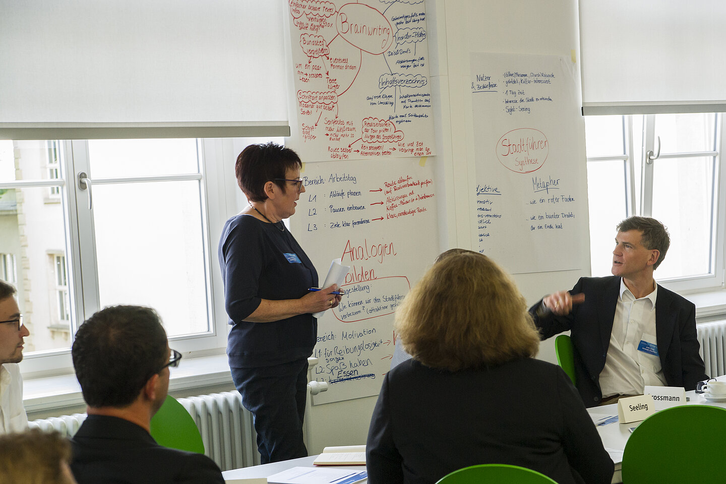 Situation aus dem Workshop. Eine Teilnehmerin steht vor einer Wand mit mehreren aufgehängten handschriftlichen Plakaten und unterhält sich mit dem Dozenten