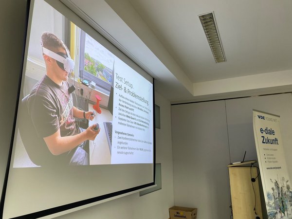 Test-Setup mit VR-Brille. (c) VDE Hochschulgruppe Leipzig Leinwand zeigt Präsenatation mit Bild einer Person mit VR-Brille am Schreibtisch sitzend