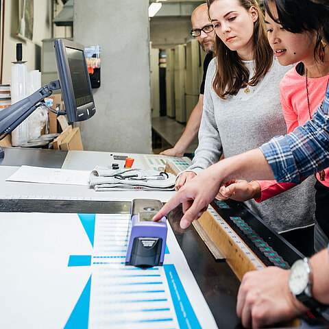 Drei Menschen beugen sich über eine Maschine und bekommen etwas erklärt Drei Menschen beugen sich über eine Maschine und bekommen etwas erklärt