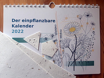 Kleiner rechteckiger Monats-Wandkalender. Links die Aufschrift "Der einpflanzbare Kalender 2022". Auf der rechten Seite des Titelblatts ein Blumenbild. Unten links ist Papier mit integrierten Blumensamen abgebildet.