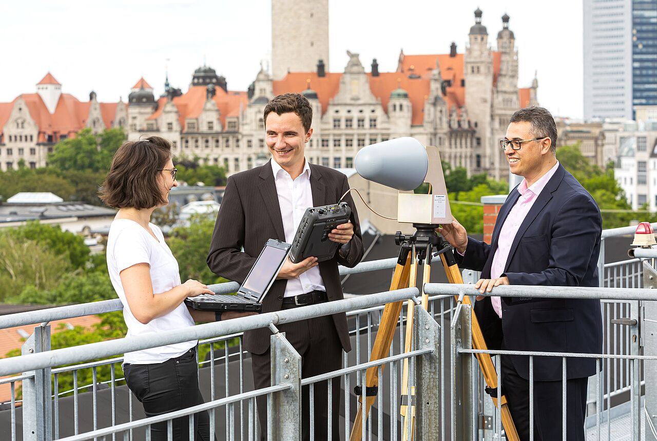 drei Personen messen etwas auf einem Dach, im Hintergrund das Neue Rathaus Leipzig
