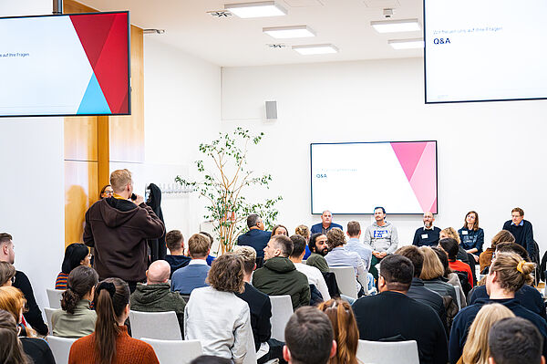 Publikum von hinten, ein Student steht und stellt eine Frage an das Plenum, das aus 7 sitzenden Personen besteht