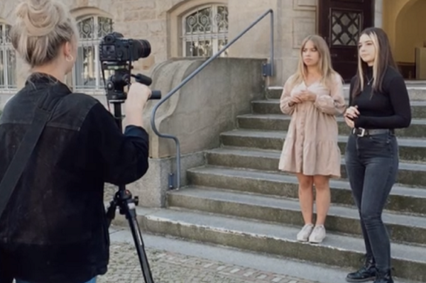 zwei Frauen im Hintergrund, im Vordergrund links Rückansicht einer Frau, die die beiden anderen mit einer Kamera auf Stativ filmt 