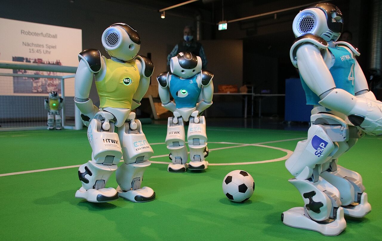 drei Roboterfußballer auf einem Kunstrasenspielfeld in einem Raum, ein Ball liegt in der Mitte von ihnen