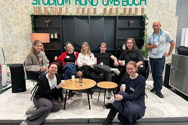 Studierende und Mitarbeiter der HTWK sitzen und posieren freudig auf der Bühne des Messestandes. 