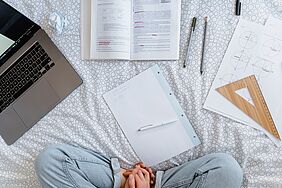 Person sitzt auf dem Bett, links ein Laptop, in der Mitte ein Buch, ein Notizblatt und rechts geometrische Zeichnungen