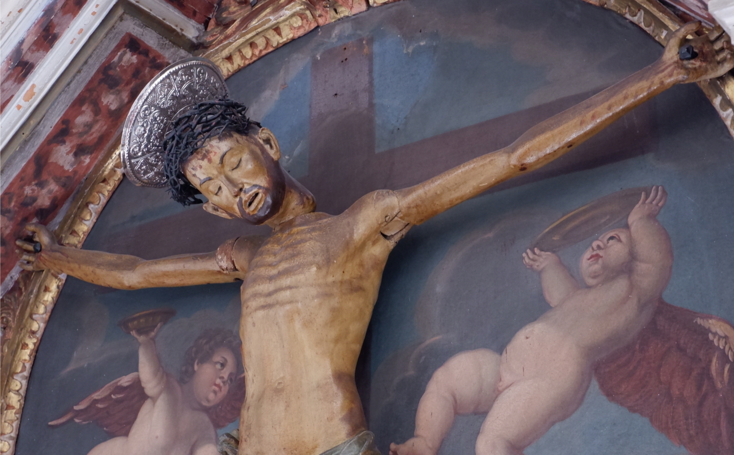 Kruzifix der Benediktinerinnen von Rab Holzfigur des gekreuzigten Christus mit beweglichen Armen und Kopf, vor einem Gemälde mit zwei Engeln und einem Kreuz