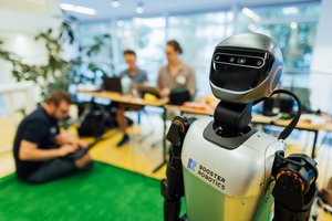 Roboter im Vordergrund, dahinter sitzen Menschen auf Kunstrasen mit Laptops