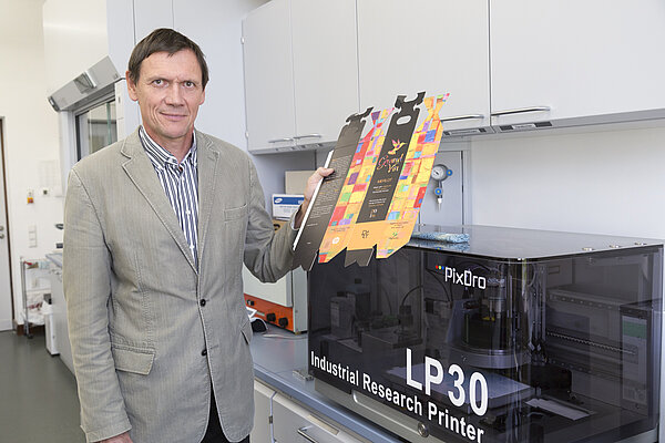 Prof. Eugen Herzau (Foto: Andreas Schröder/HTWK Leipzig) Prof. Eugen Herzau steht neben einer für Forschungszwecke optimierten Digitaldruckmaschine. In der Hand hält er einen digital bedruckten Weinkarton.