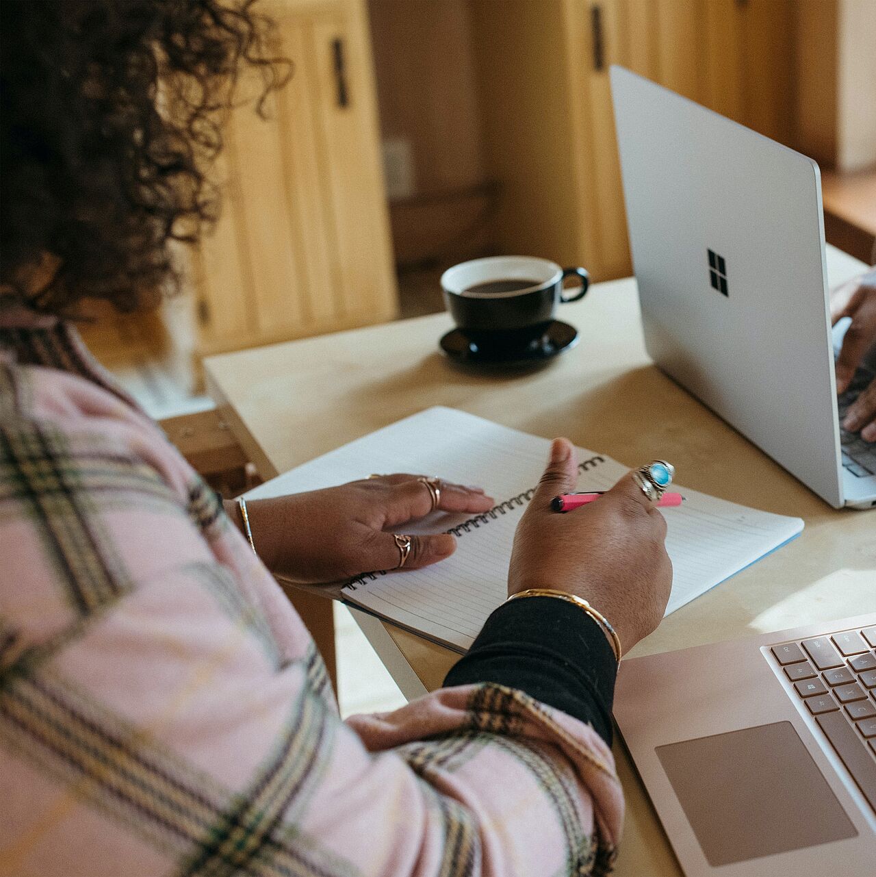 Bild zeigt eine Frau am Schreibtisch wie sie etwas in ein Heft schreibt und zwei Laptops.