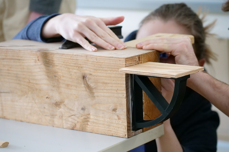 Nahaufnahme eines dunklen Elementes mit schmaler Regalauflage an einem Holzblock befestigt