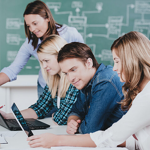 Bild zeigt 4 Personen an einem Tisch mit Laptops und einer Tafel im Hintergrund.