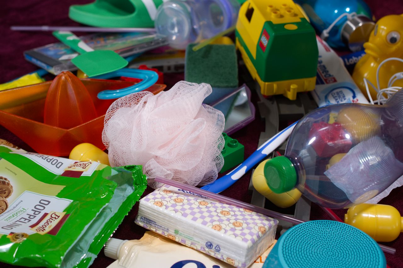Auf einer dunklen Unterlage liegt eine große Ansammlung verschiedener Alltags- und Haushaltsgegenstände. Zu sehen sind unter anderem mehrere Kunststoffflaschen und -verpackungen, eine Zahnbürste, Taschentücher, ein rosa Duschschwamm, ein grüner Reinigungsschwamm, Spielzeug und eine gelbe Spielzeugente. Die Gegenstände liegen ungeordnet übereinander und füllen nahezu das gesamte Bild aus. Viele der sichtbaren Objekte bestehen aus Kunststoff.