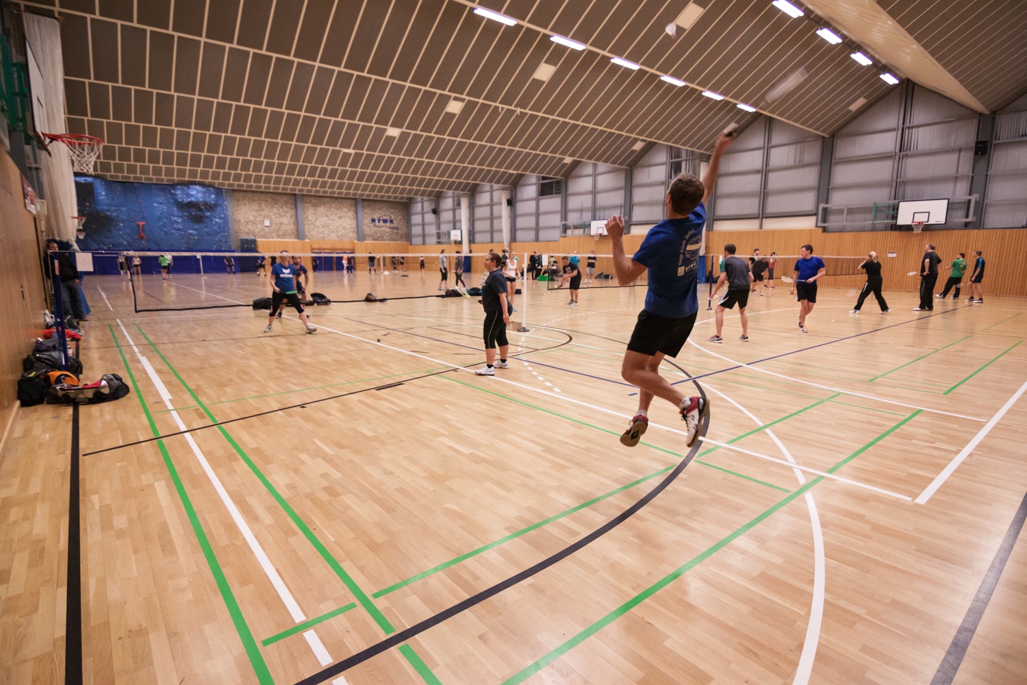 In einer Sporthalle spielen auf mehrenen Feldern Studenten Badminton. Im Vordergrund springt ein Spieler in die Luft um den Badmintonball zu treffen.