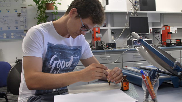 Schnittbild eines Studenten im Labor. Er sitzt an einem Tisch und bearbeitet Druckvorlagen.