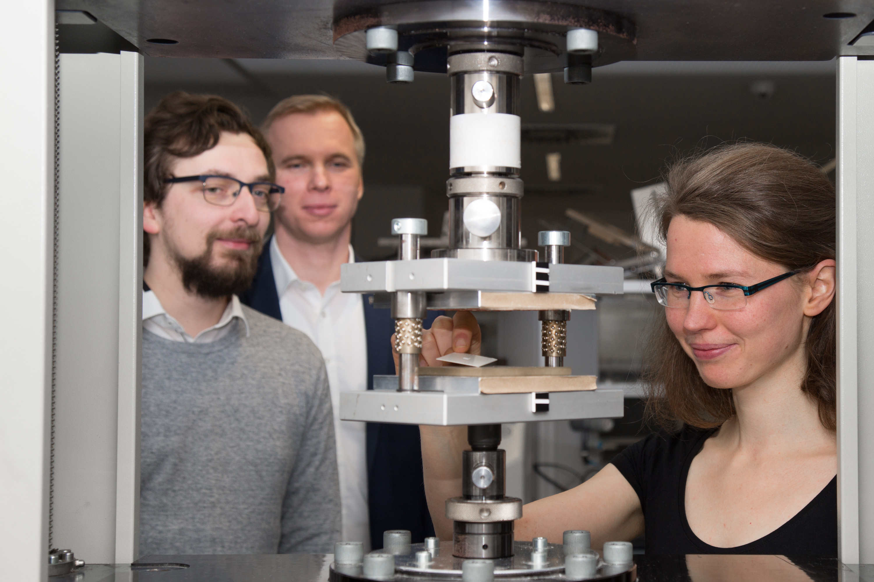 Darstellung einer Zwick Universalprüfmaschine mit Prägewerkzeug. Dies sind parallel geführte Platten, zwischen denen sich die Patritze und Matritze befinden. Gezeigt ist beispielhaft eine geprägte Probe, die gerade aus dem Werkzeug entnommen wird. Auf dem Bild sind gezeigt: Prof. Stephan Schönfelder, Florian Wallburg und Ulrike Käppeler.